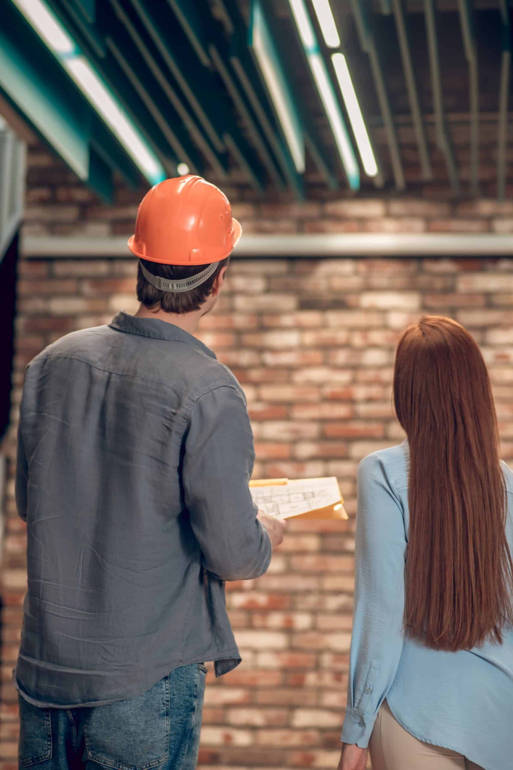 A rear view of a construction worker wearing an orange hard hat and holding a clipboard, standing with a woman looking toward an exposed brick wall inside a building.